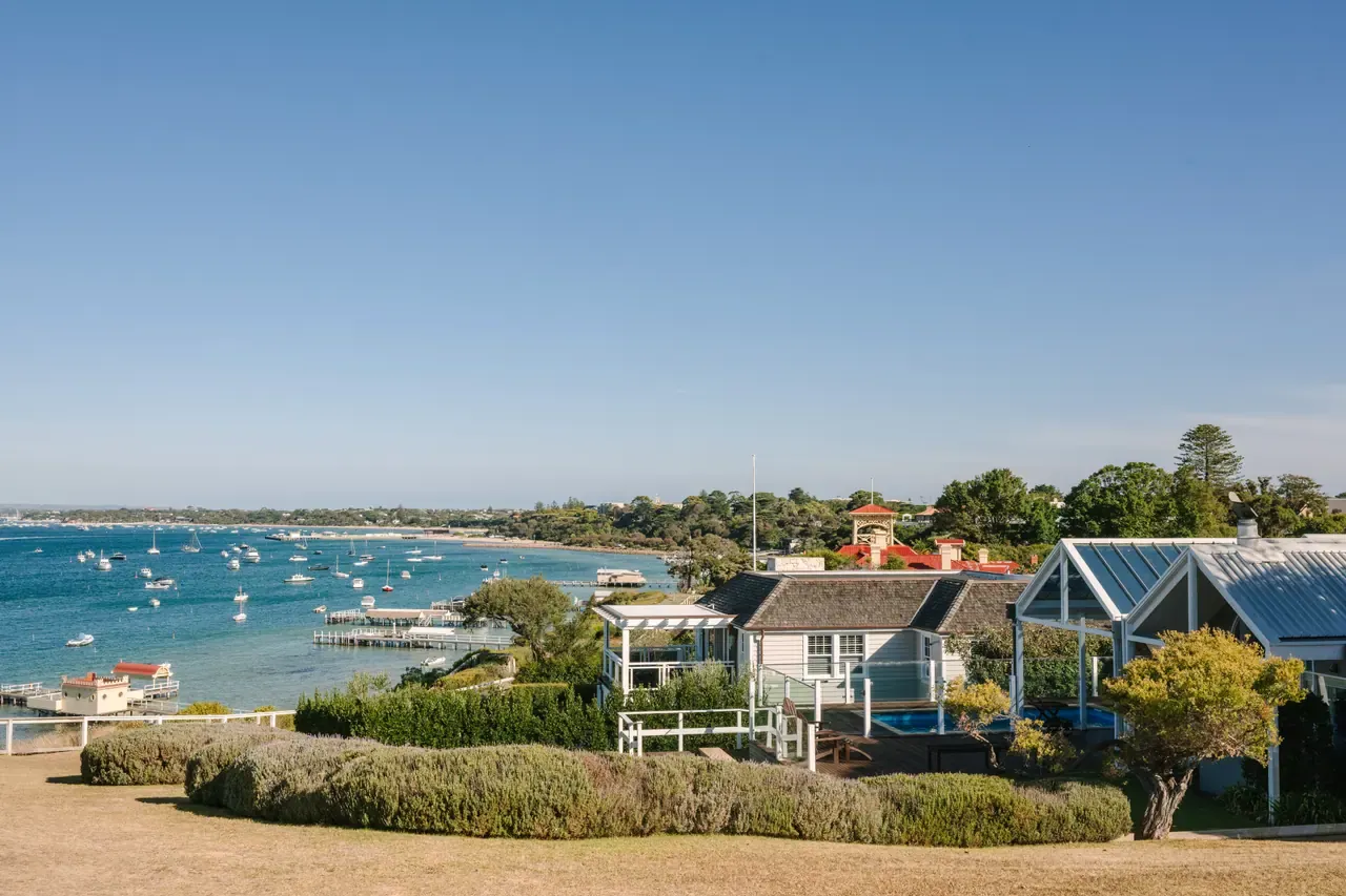 Clifftop lawn and weatherboard houses above a bay dotted with moored boats and jetties along Millionaire's Walk