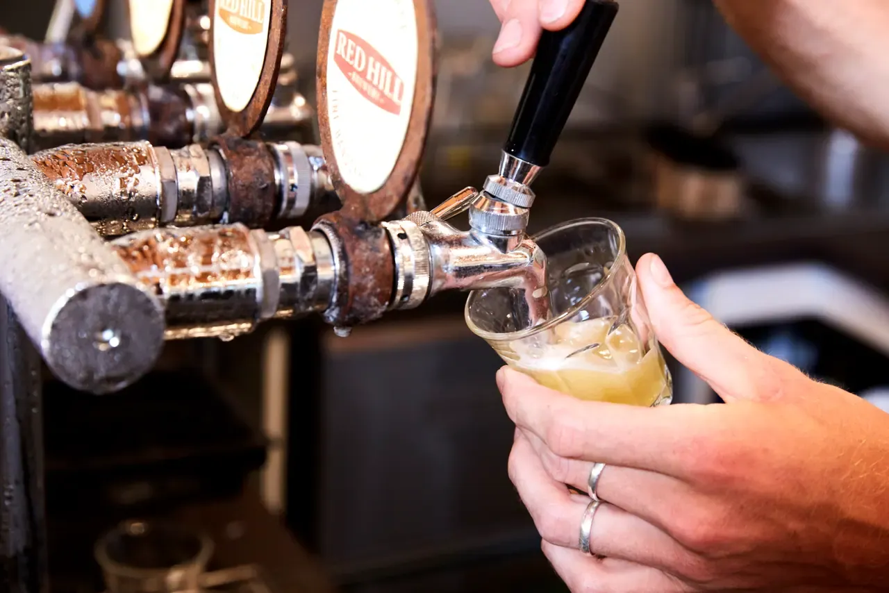 A hand pulls a pale beer into a small glass from a row of worn metal taps with a Red Hill Brewery tap badge