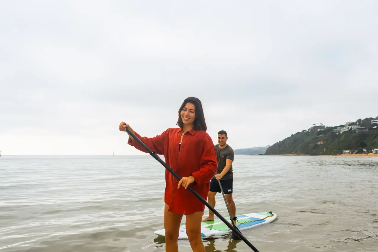 A smiling paddleboarder in a red shirt, followed by a second paddler, on calm water off Mills Beach