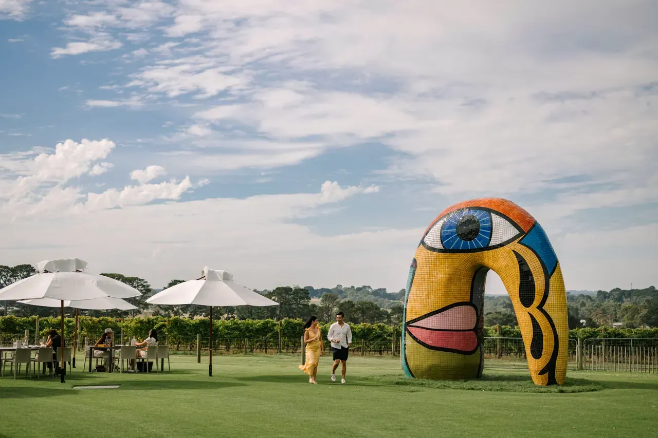 Diners sit under white umbrellas on the lawn at Pt. Leo Estate beside a large mosaic face sculpture, under a wide cloudy sky