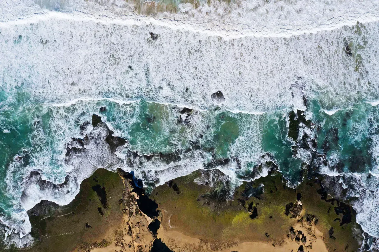 Overhead view of white surf breaking over dark reef and rock shelf at Sorrento Back Beach, with turquoise water between the rocks