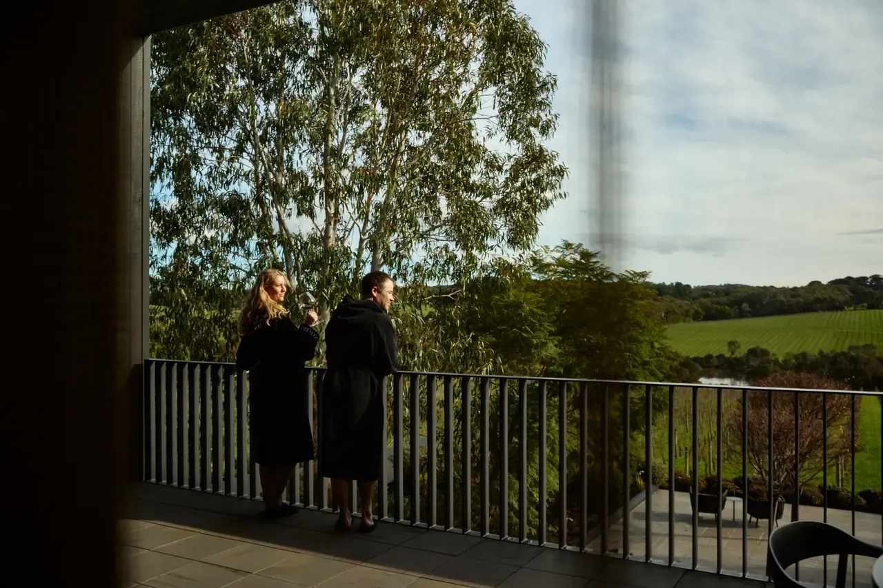 Two guests in dark robes stand on a balcony at Jackalope Hotel, looking out over gum trees to vineyard rows
