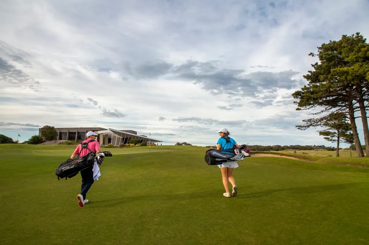 Two golfers carry their bags across a fairway towards a modern clubhouse under a broad, cloudy sky.