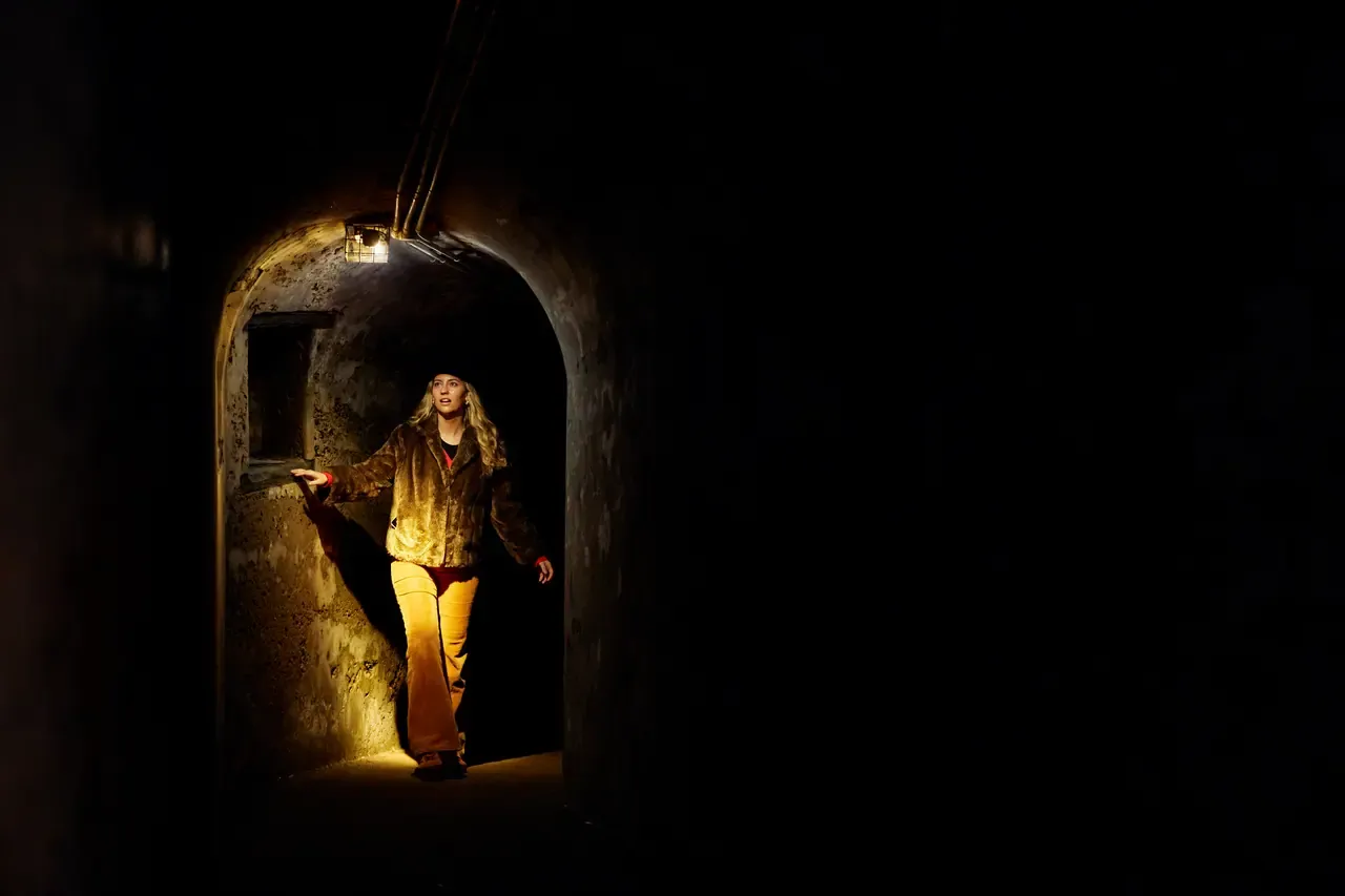 A person walks through a dim concrete tunnel at the Point Nepean fortifications, lit by a single caged lamp overhead
