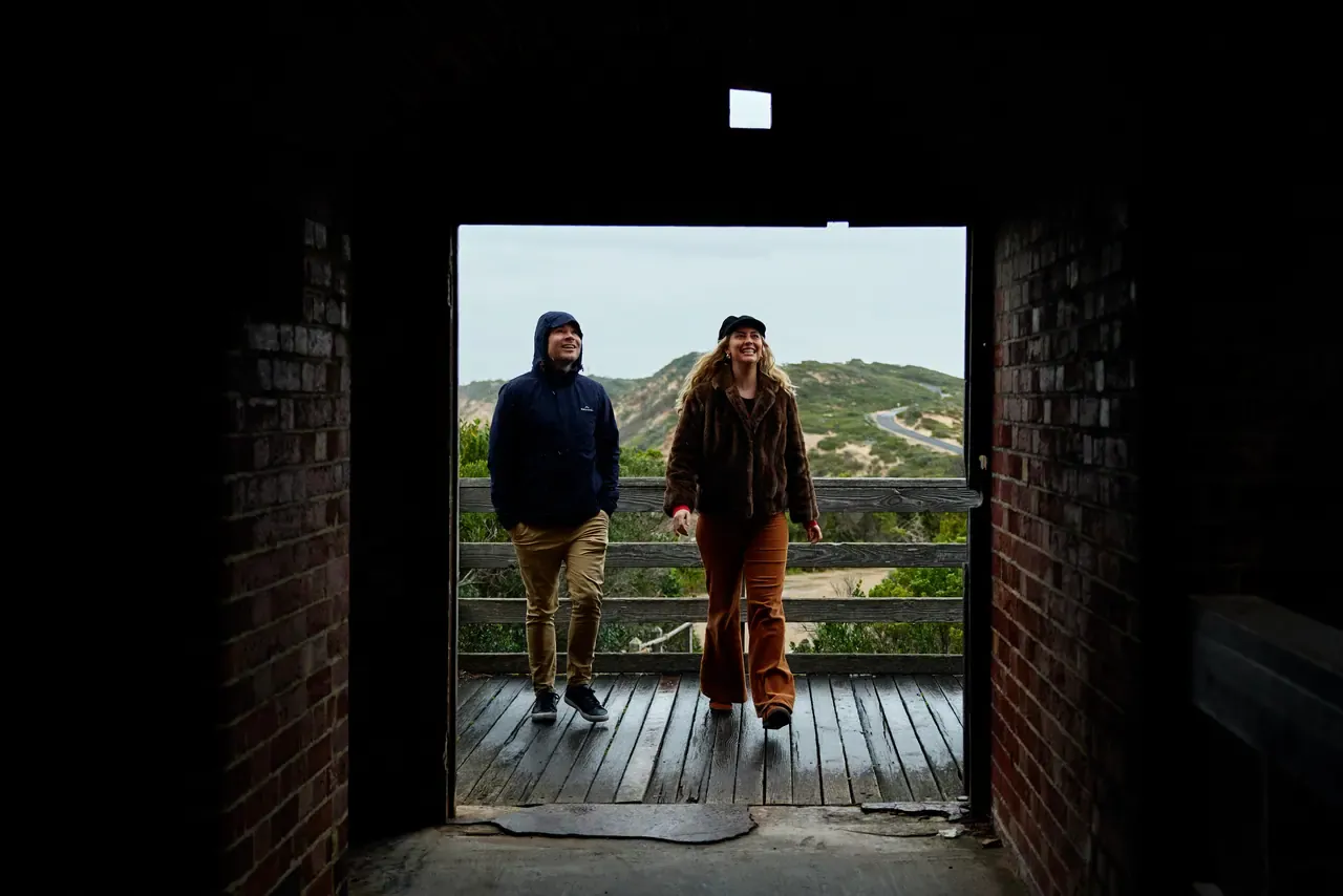 Two people step from a wooden deck into a dark brick doorway at Point Nepean, with scrubby hills and a winding road behind