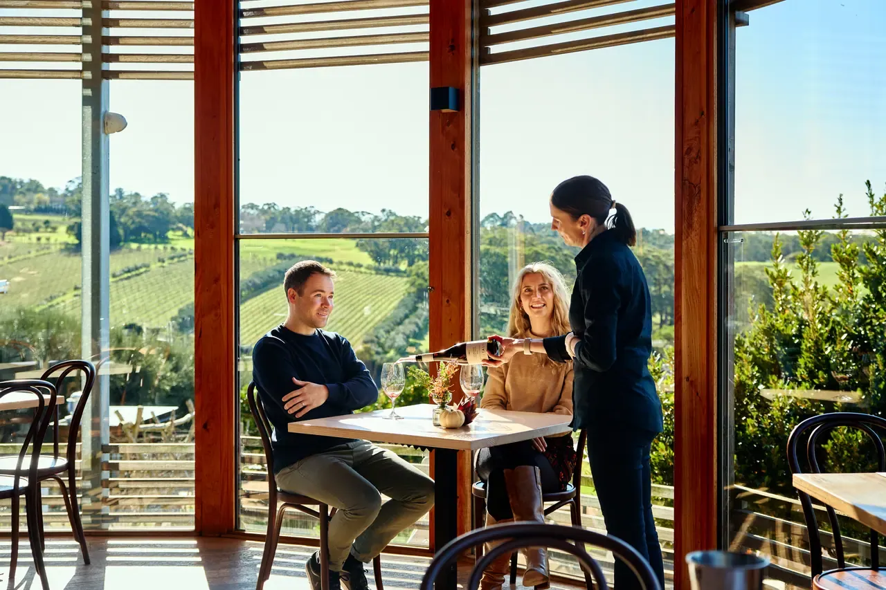 Server pouring red wine for a couple at a table in the glass-walled Montalto dining room, with vineyard views behind