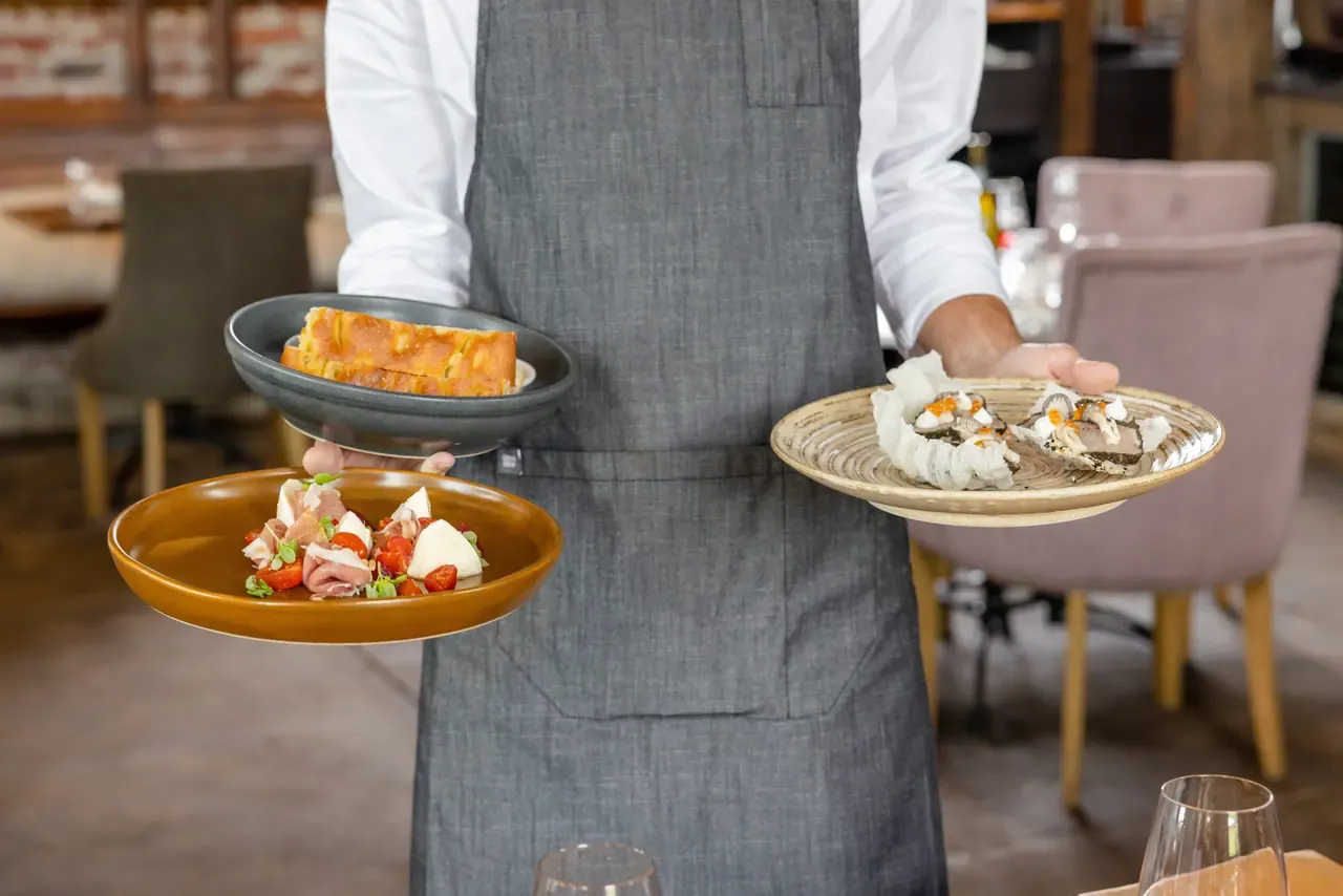 A server in a grey apron carrying three plates of shared dishes, including bread and oysters, through the Trofeo Estate dining room