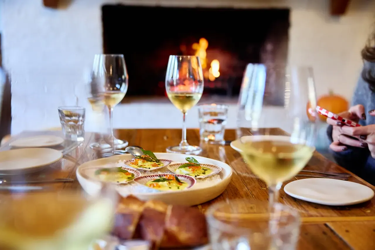 Scallops on the half shell served on salt with bread and glasses of white wine, an open fire glowing behind