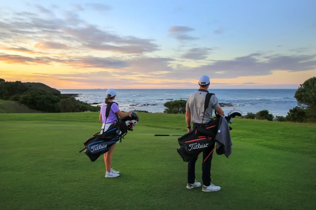 Two golfers carrying bags look out over the ocean from the course at Flinders under a colourful sunset sky.