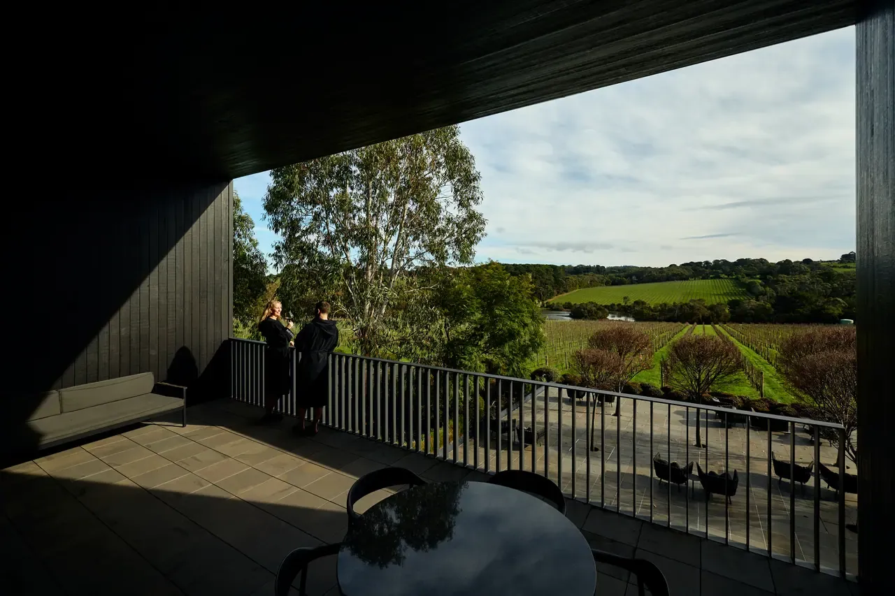 Two guests in robes on a shaded terrace at Jackalope Hotel, looking over the vineyard and green hills under a cloudy sky