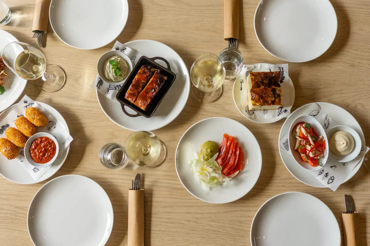 Overhead view of a timber table set with croquettes, cured salmon, glazed pork and white wine on white plates