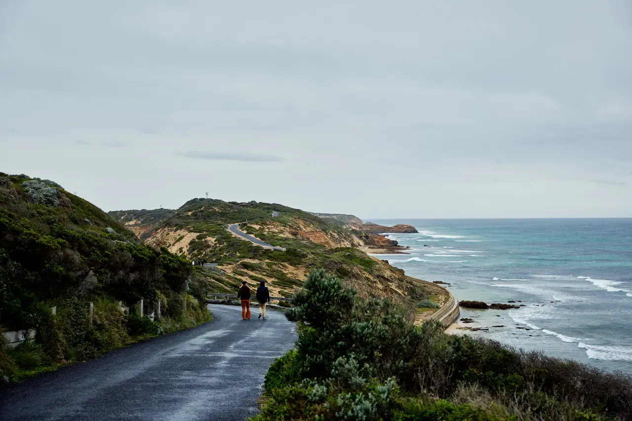 Two walkers head along a wet coastal road towards grassy headlands and surf under an overcast sky at Point Nepean National Park.