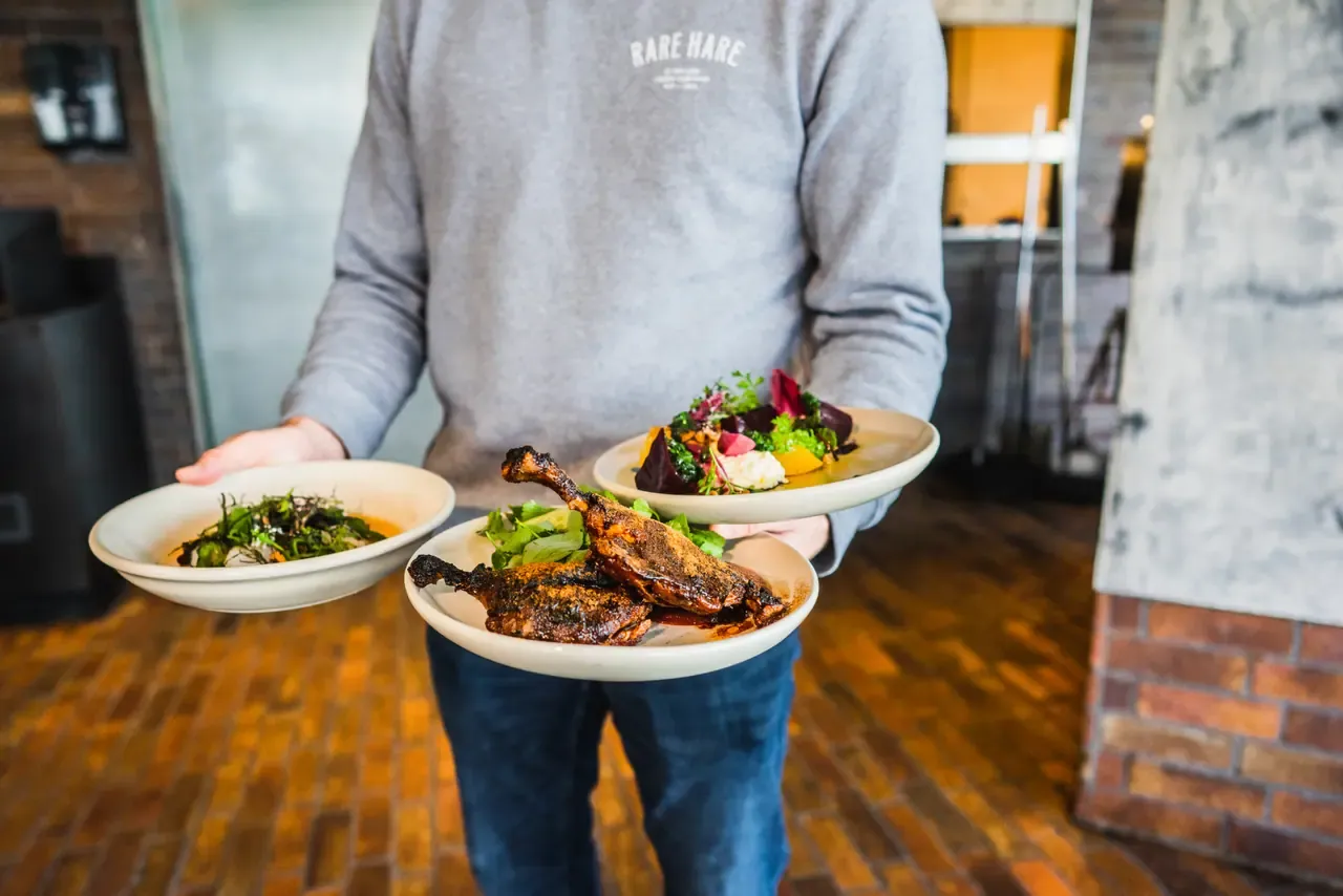 A staff member in a Rare Hare jumper carries three plates, including roast duck and a beetroot salad