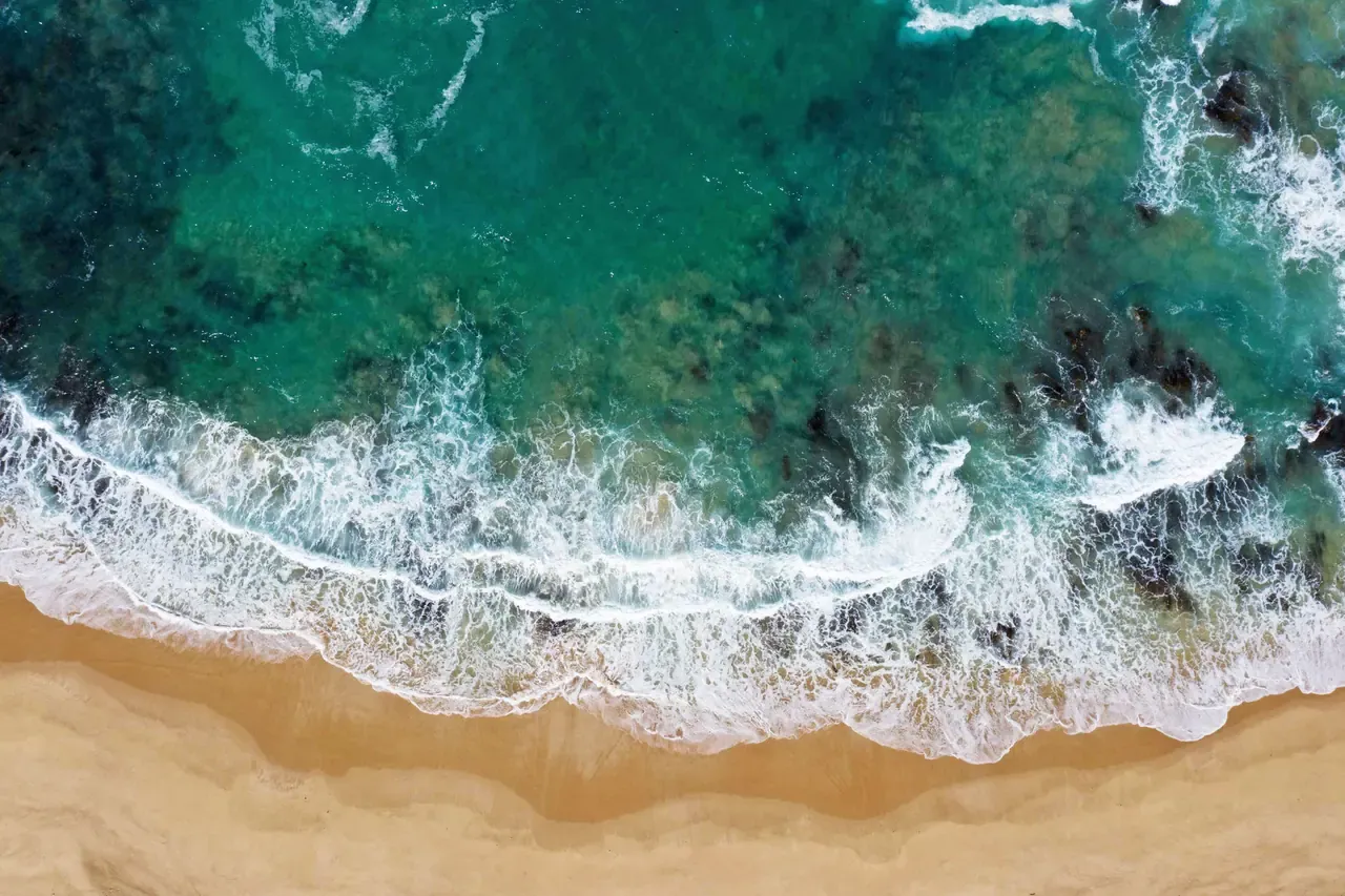 Overhead view of waves breaking over turquoise water and dark reef onto the golden sand of Sorrento Back Beach