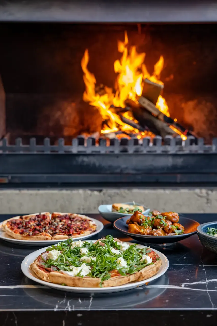 Pizzas and a bowl of glazed chicken wings on a marble bench in front of an open wood fire at St Andrews Beach Brewery