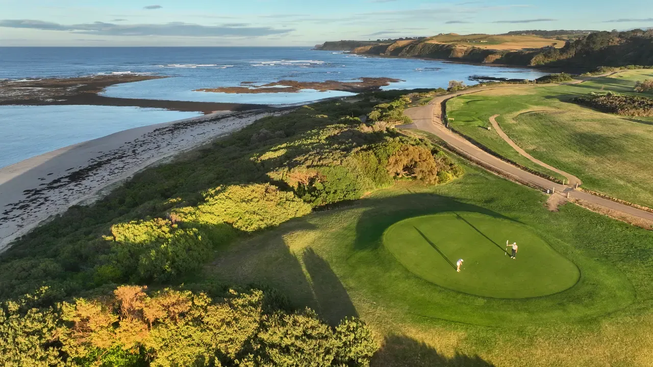 Aerial view of two golfers on a coastal green at Flinders Golf Course, with long shadows, coastal scrub and the ocean beyond in low sun.