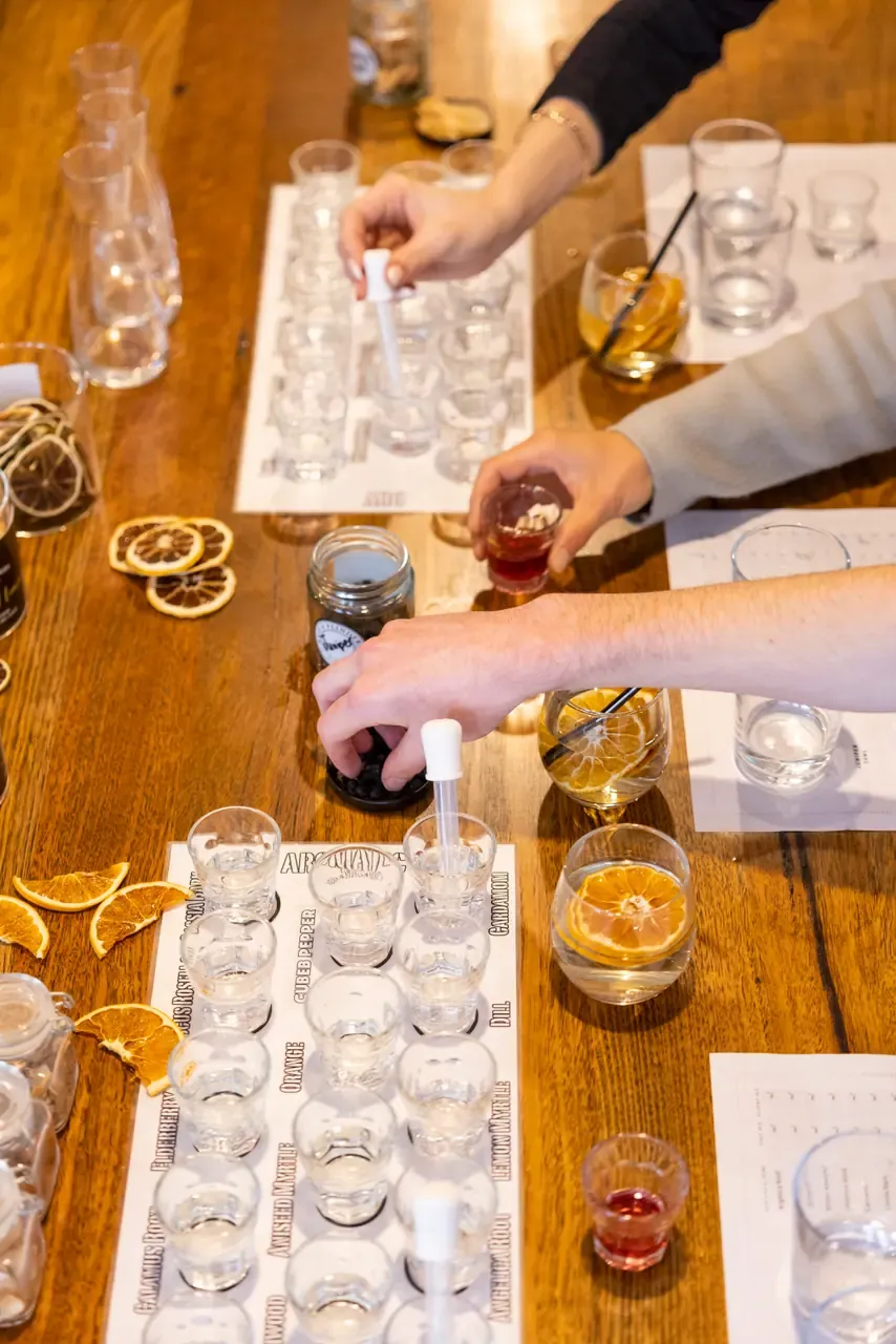Hands use droppers to blend botanicals from rows of labelled shot glasses during a gin-making class at a timber table