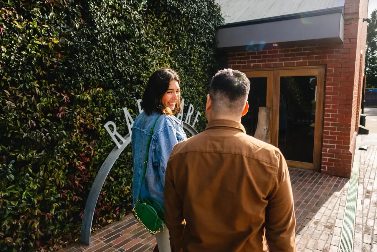Two visitors walk past the ivy wall and arched Rare Hare sign towards the red-brick entrance in sunshine