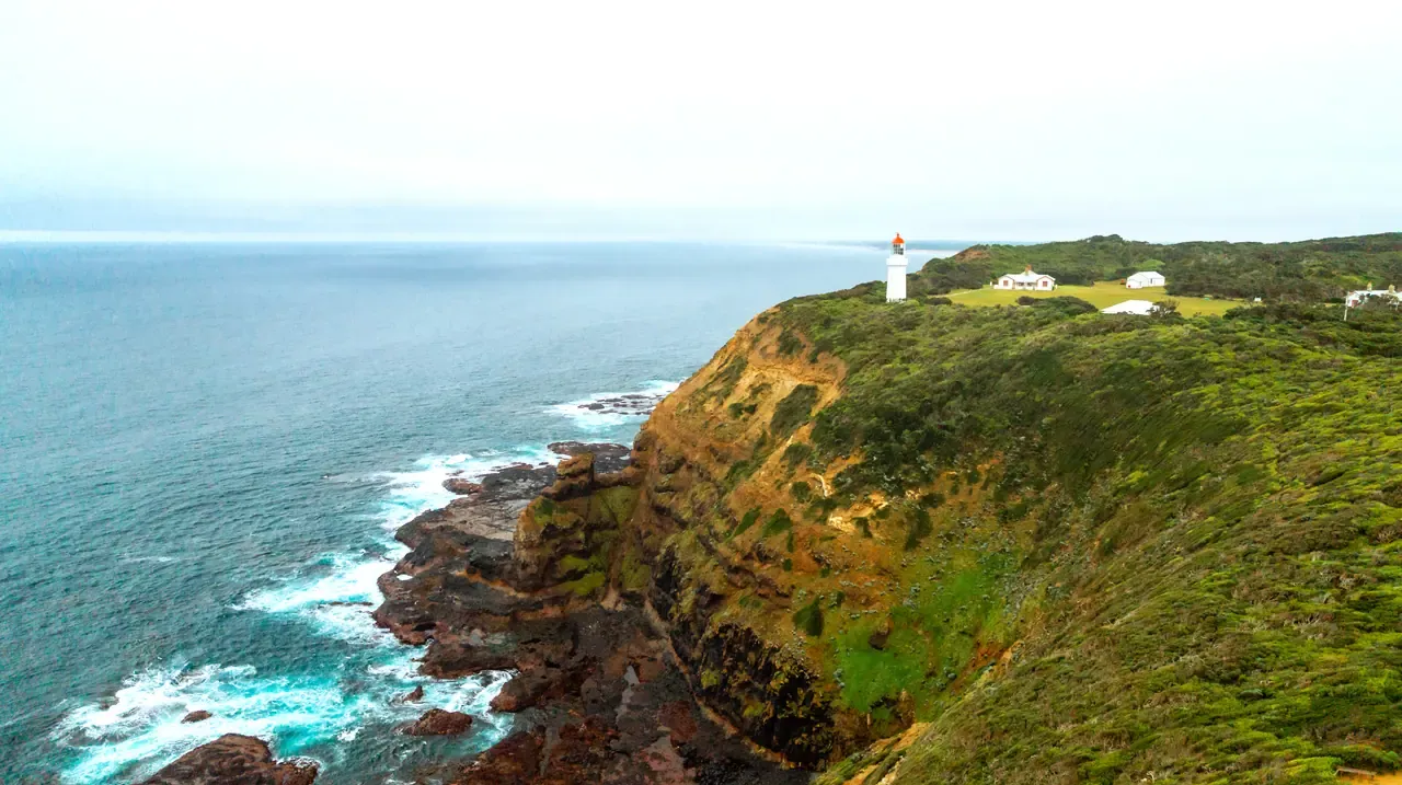 Aerial view of green cliffs dropping to a rocky shore, with the Cape Schanck lighthouse and cottages on the headland