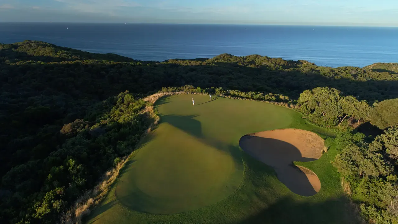 Aerial view of a green and bunker cut into dense coastal scrub at The National Golf Club, with the open sea behind