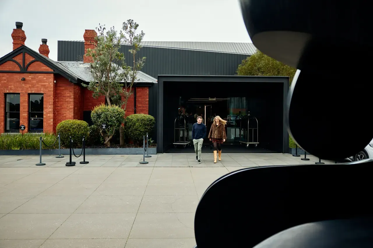 Two guests leave the black-framed Jackalope Hotel entrance beside a red-brick building in Merricks North