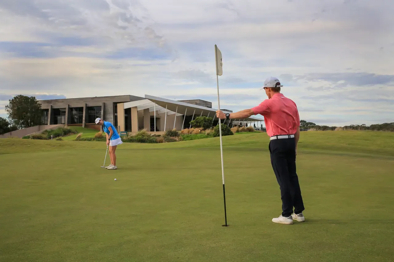 A golfer holds the flag while another putts on a green at Moonah Links, the angular clubhouse building behind