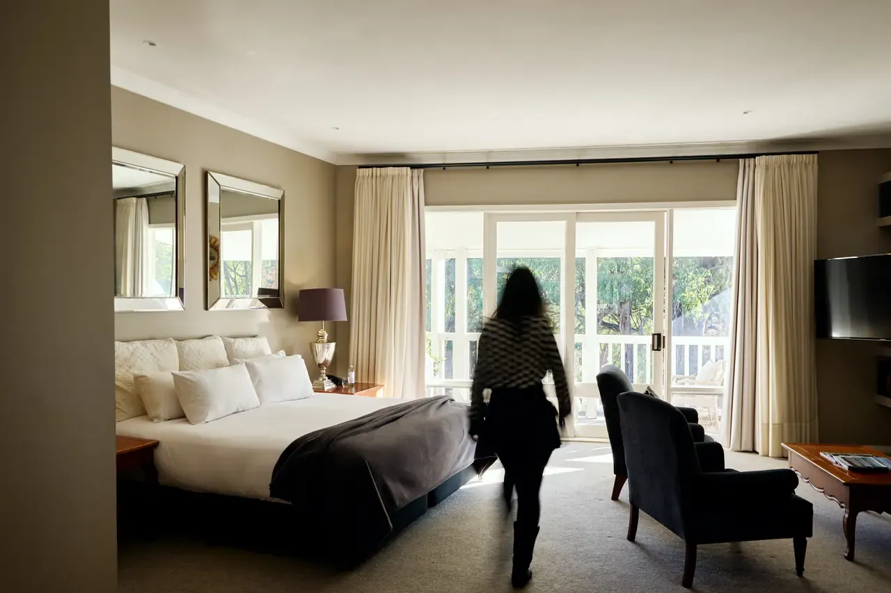 A guest walks through a bedroom at Lindenderry with a king bed, mirrors, armchairs and French doors onto a balcony