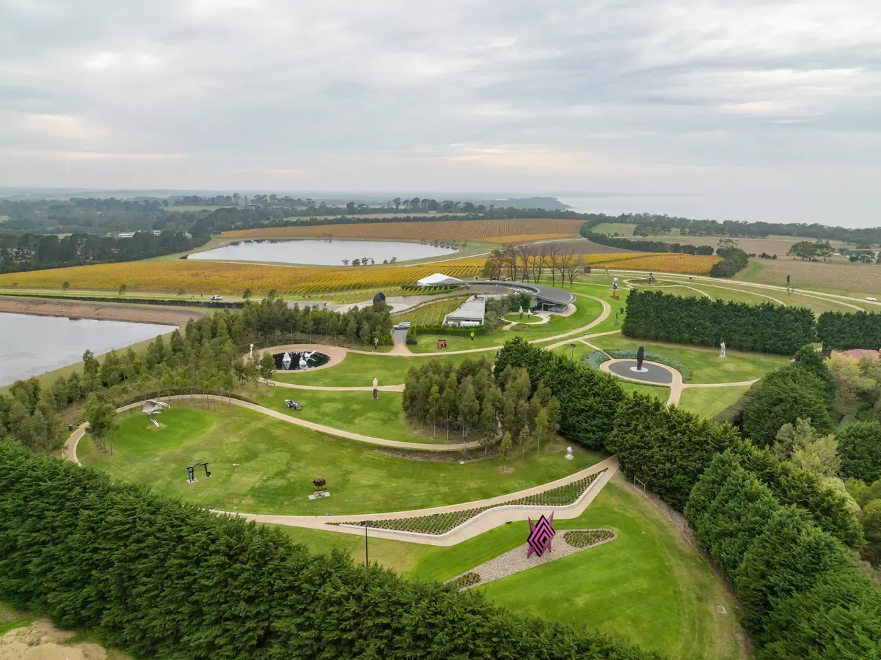 Aerial view of the Pt. Leo Estate sculpture park, with lawns, winding paths and cypress hedges above vineyards, a lake and the bay