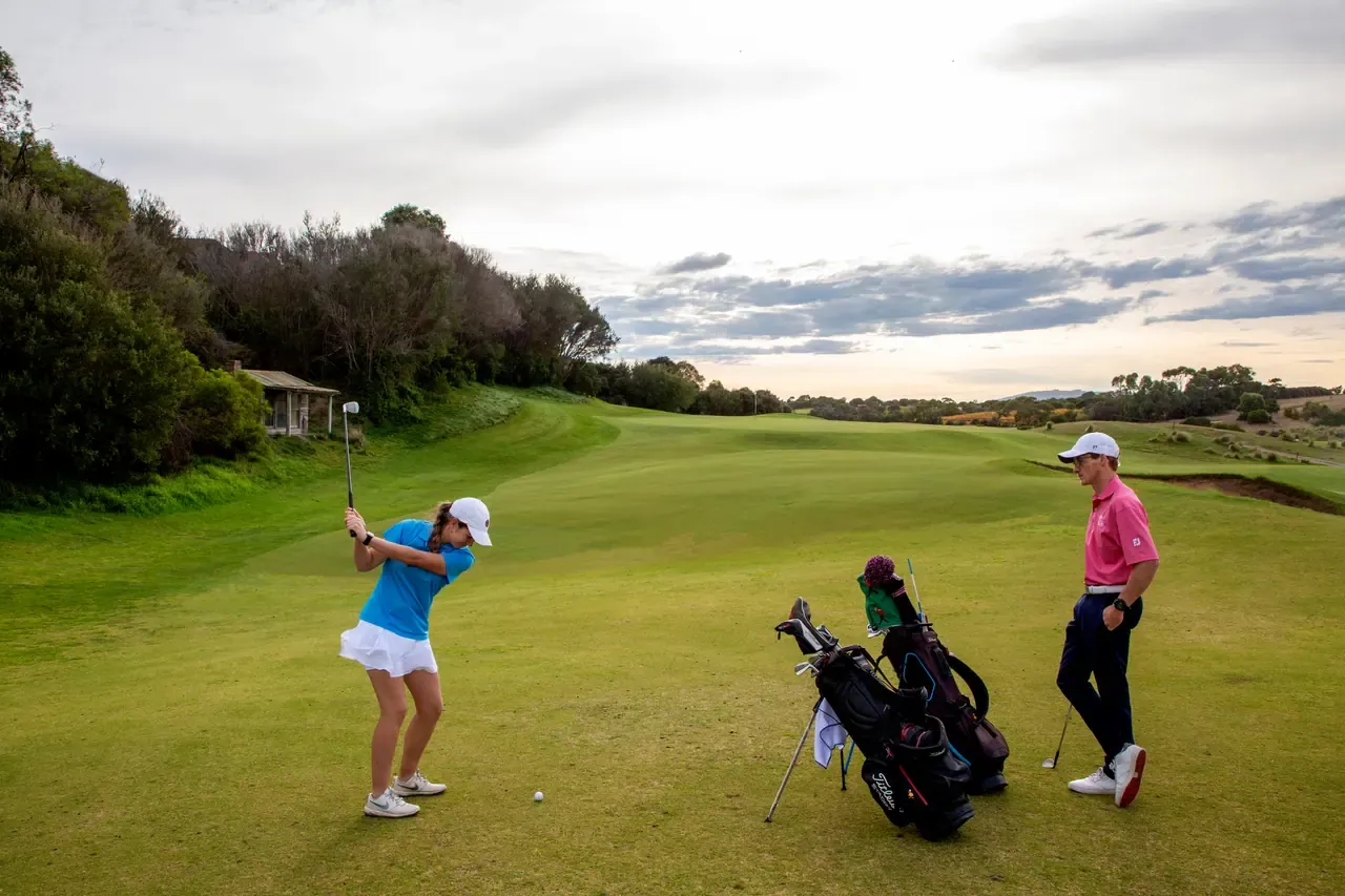 A golfer lines up a shot on a tree-lined fairway at Moonah Links while a partner stands by the golf bags