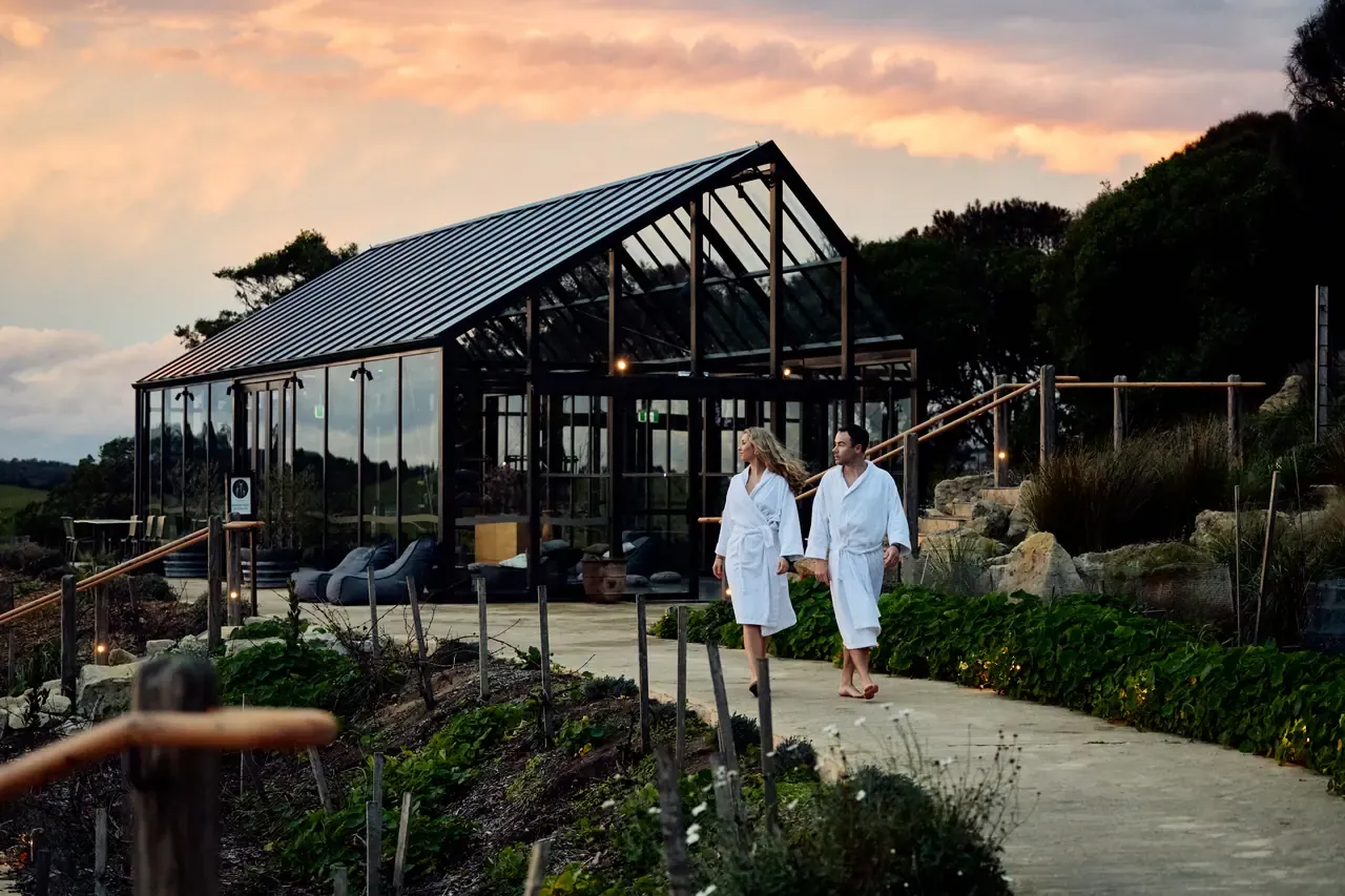 Two guests in white robes walk along a path past a glass pavilion under a pink dusk sky at Peninsula Hot Springs