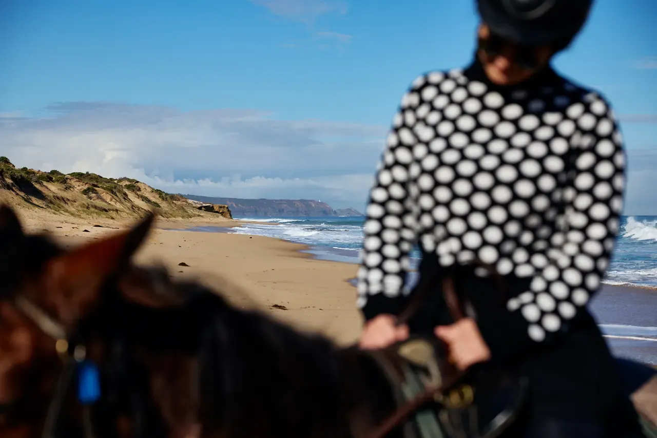 Close view of a rider in a spotted jumper on horseback, blurred, with the dunes and surf of Gunnamatta Beach in sharp focus behind