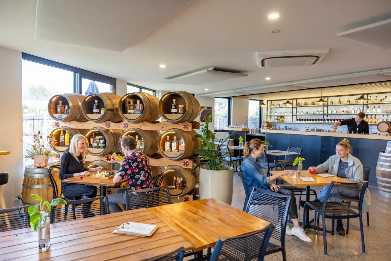 Guests play chess and share drinks at tables in the Bass and Flinders Distillery bar, beside a wall of bottle-filled barrels