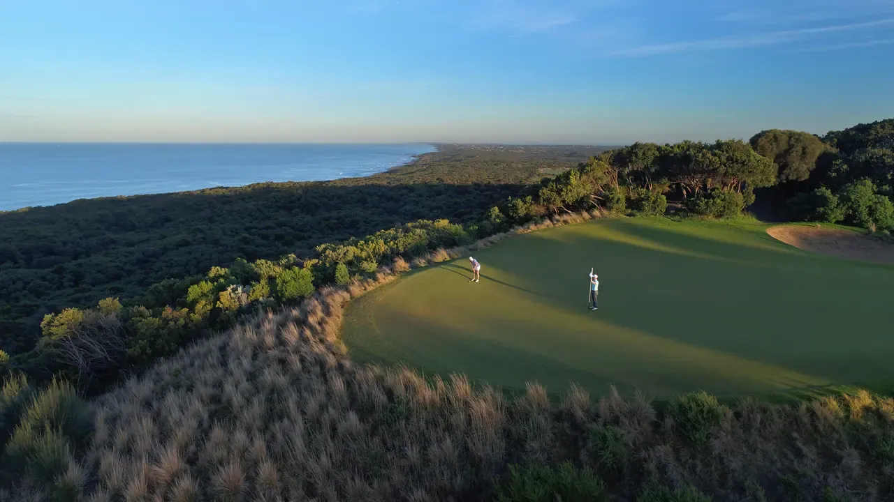 Aerial view of golfers on a green at The National Golf Club, edged by tussock and scrub, with the coastline stretching away