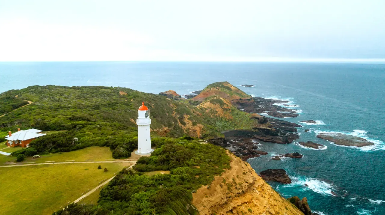 Aerial view of the Cape Schanck lighthouse on a scrubby clifftop, with rock reefs and surf stretching into the ocean