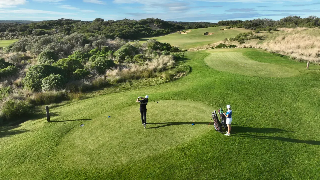 Elevated view of a golfer driving from a tee at St Andrews Beach Golf Course, a partner by the bags and the green ahead in the dunes