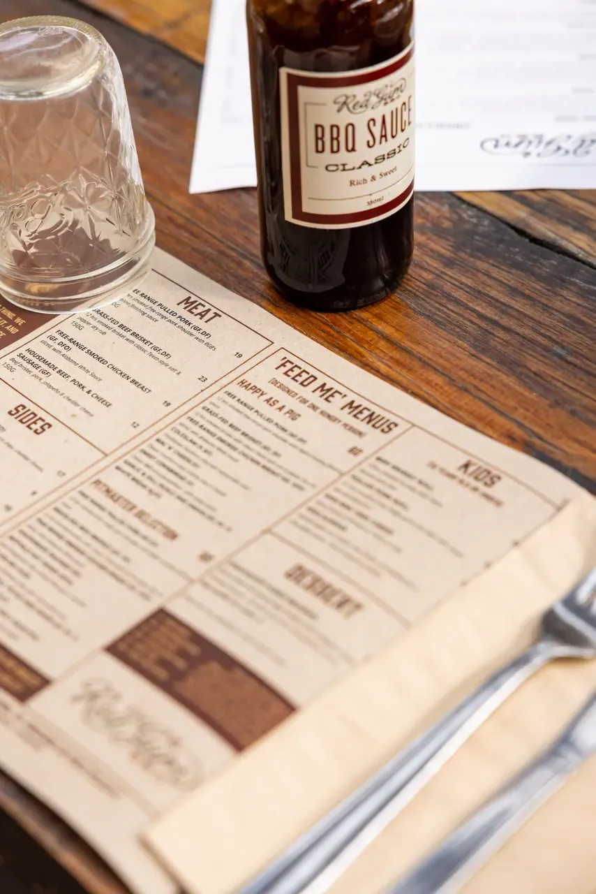 A bottle of house barbecue sauce and an upturned glass sit on a paper menu beside cutlery on a timber table