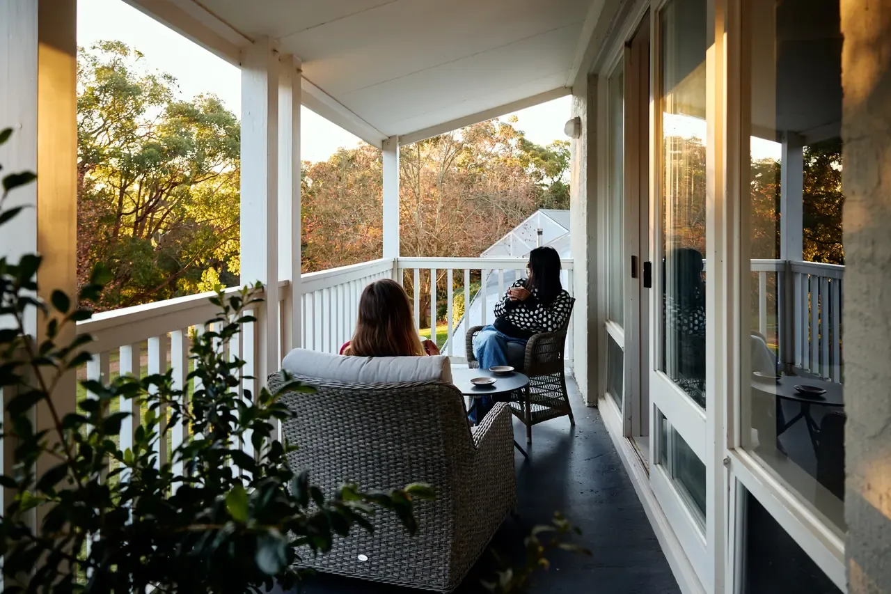 Two guests sit in wicker chairs on a white-railed upstairs verandah looking out over gum trees in the late afternoon light
