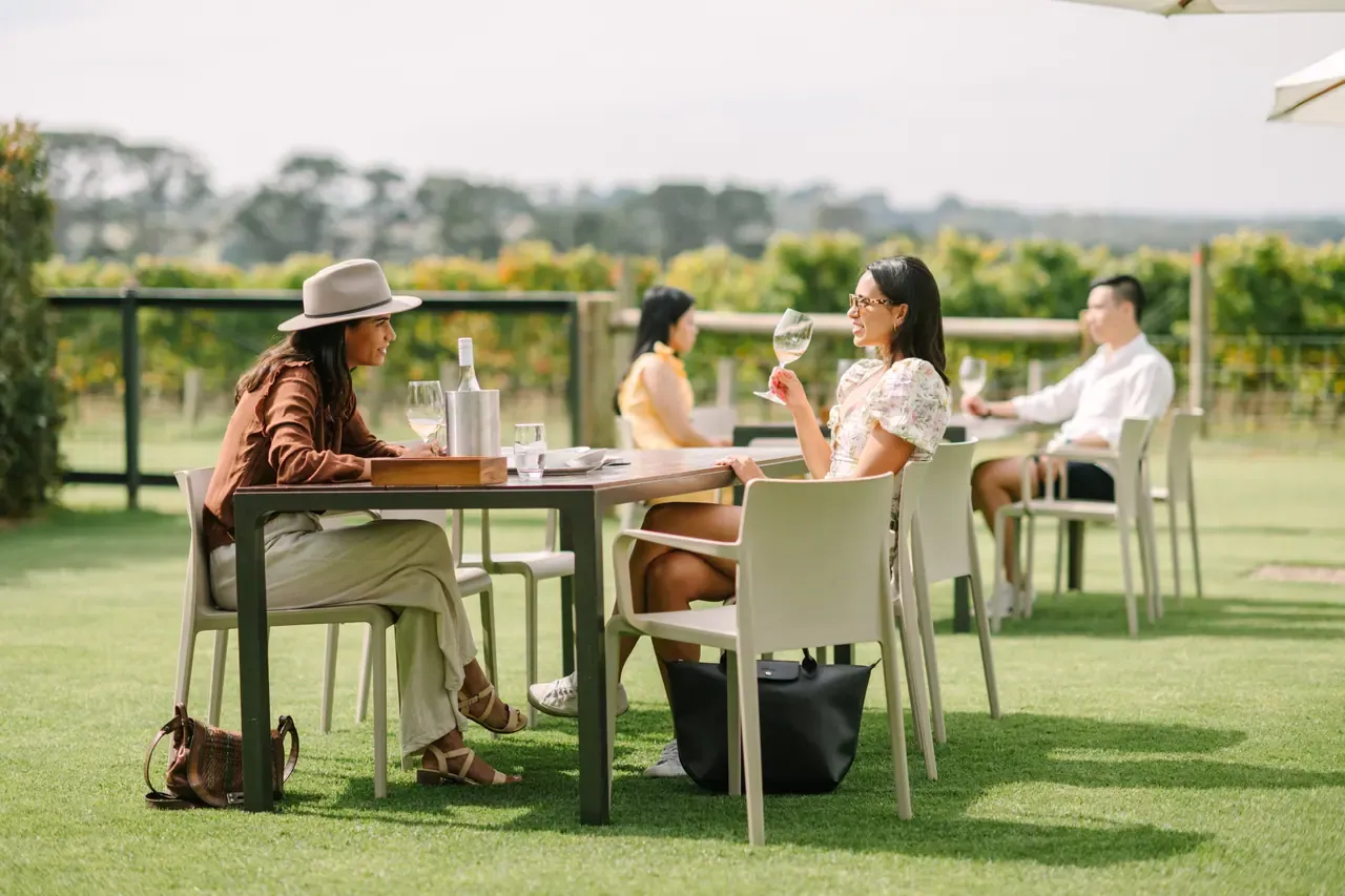 Guests share wine at an outdoor table on the lawn at Pt. Leo Estate, with a vineyard and tree-lined hills behind
