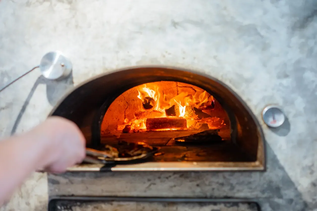 Logs burn inside a domed wood-fired oven as a hand reaches in with tongs, seen from the front of the oven
