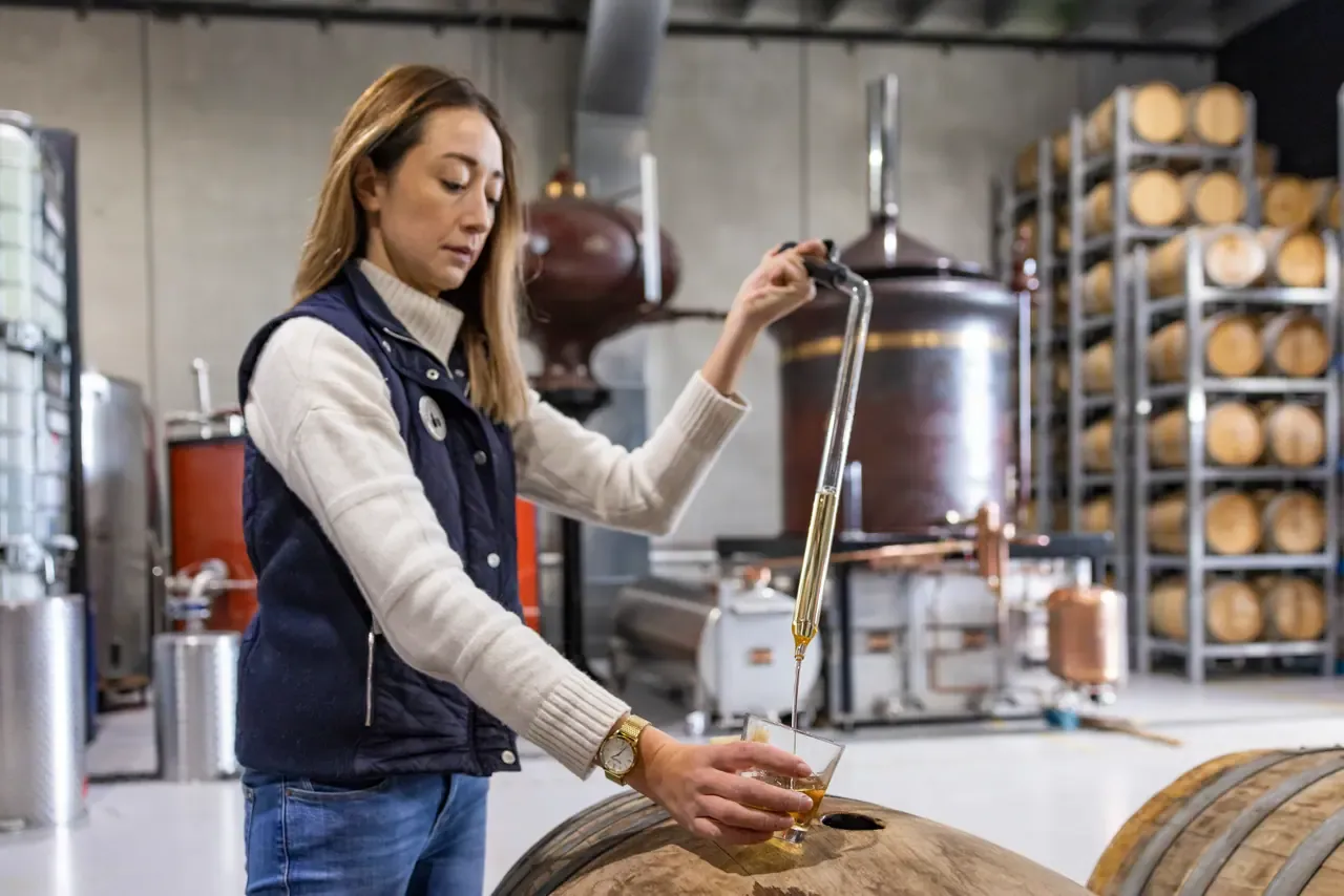 A distiller draws spirit from an oak barrel with a glass thief, in front of a copper still and racks of barrels