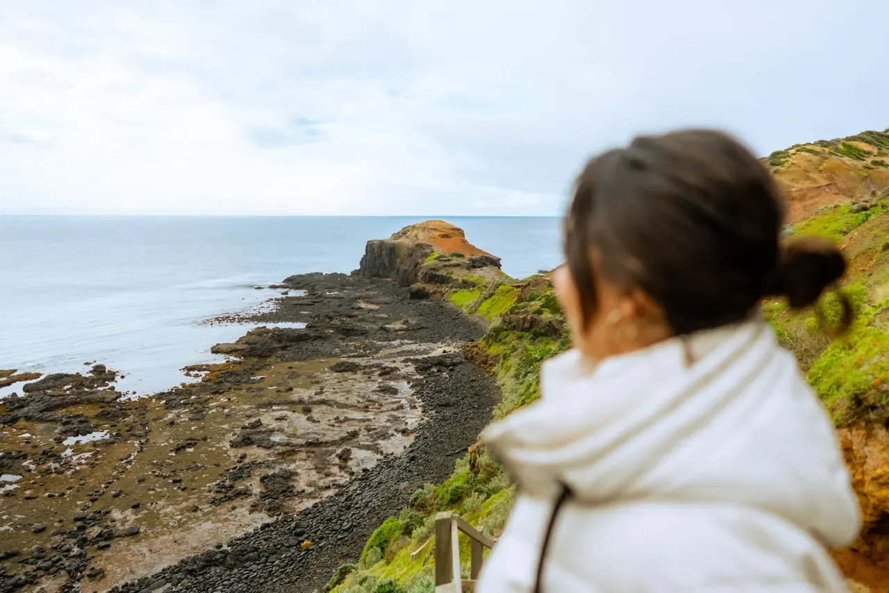 A walker in a white jacket looks out over the rock platform and headland below the Cape Schanck Boardwalk