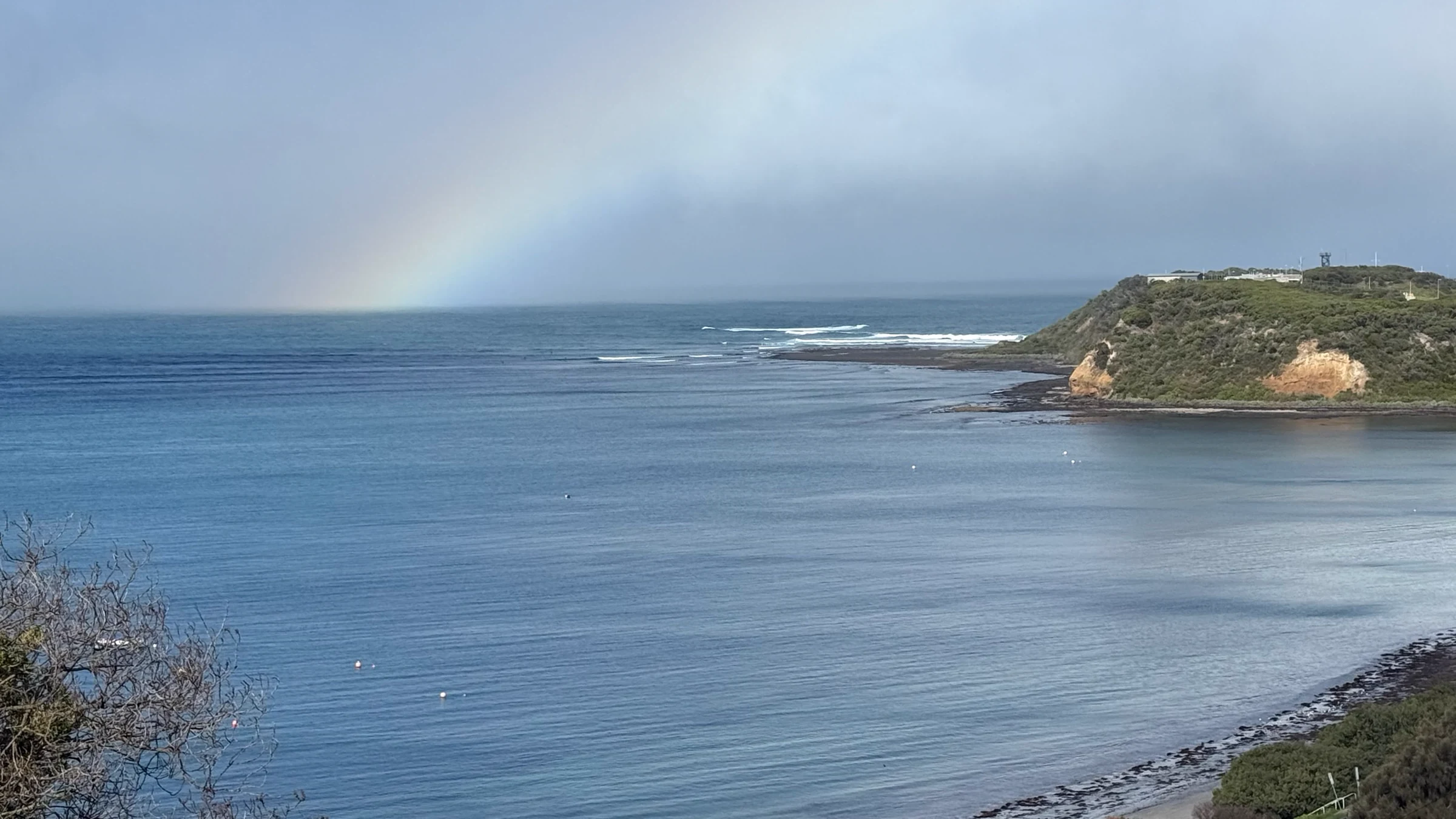 Rain squall clearing over Cape Schanck bay in winter, rainbow arcing over mist-softened headland and steel-grey Bass Strait