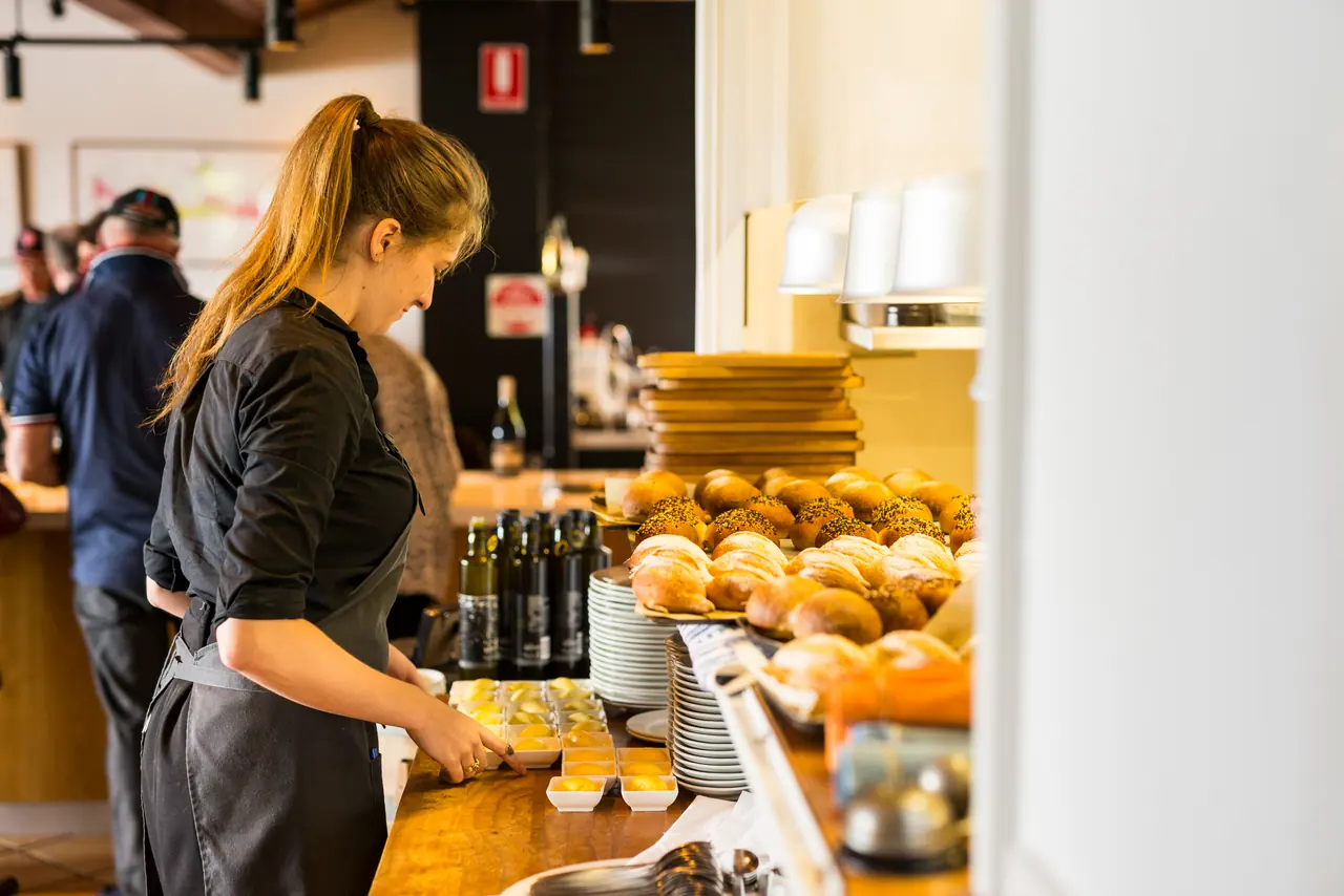 A staff member preparing small dishes of butter beside trays of bread rolls at a restaurant service counter