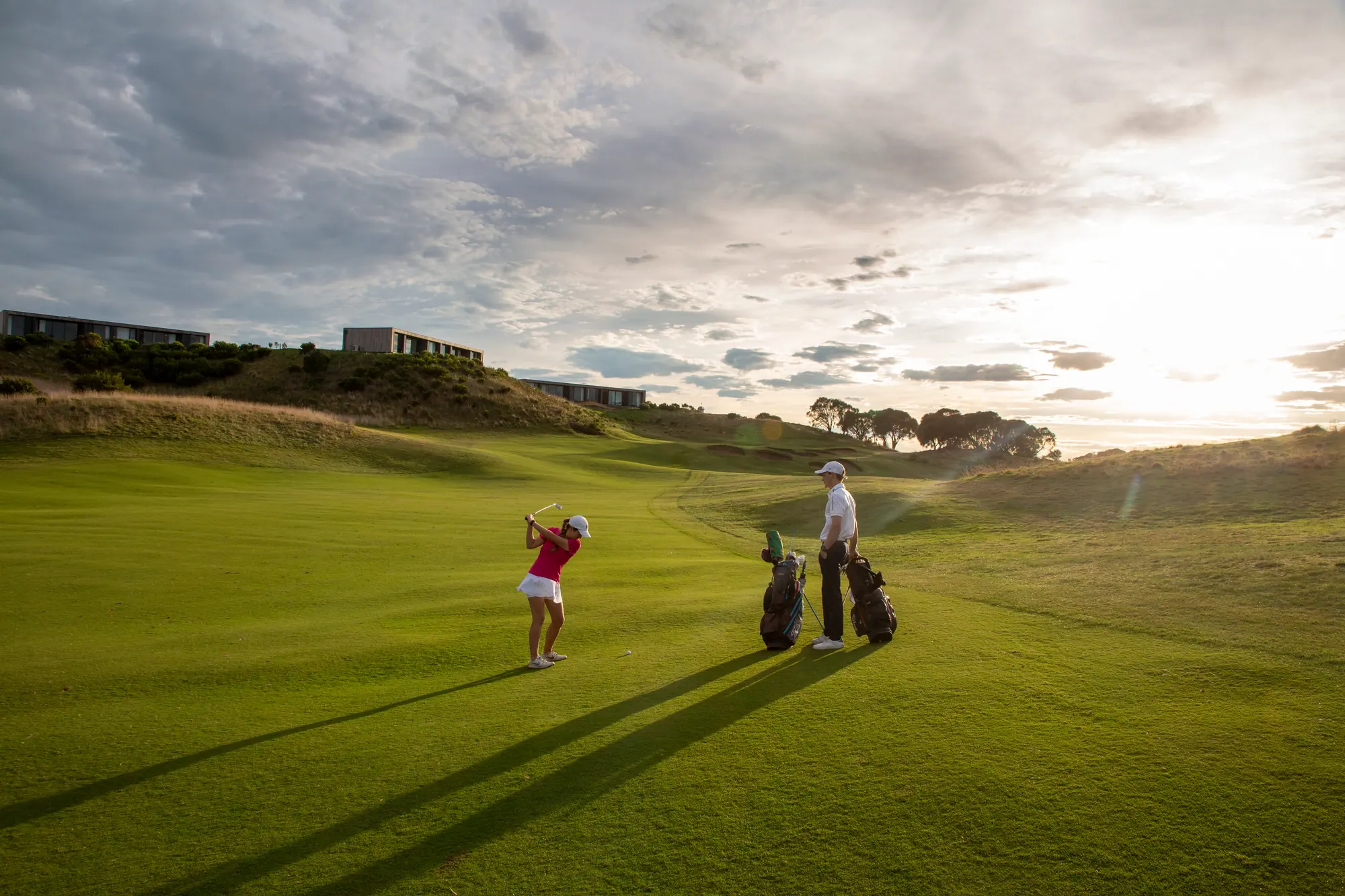 A golfer plays a fairway shot at The Dunes Golf Links as a partner stands by the bags, long shadows under a bright cloudy sky