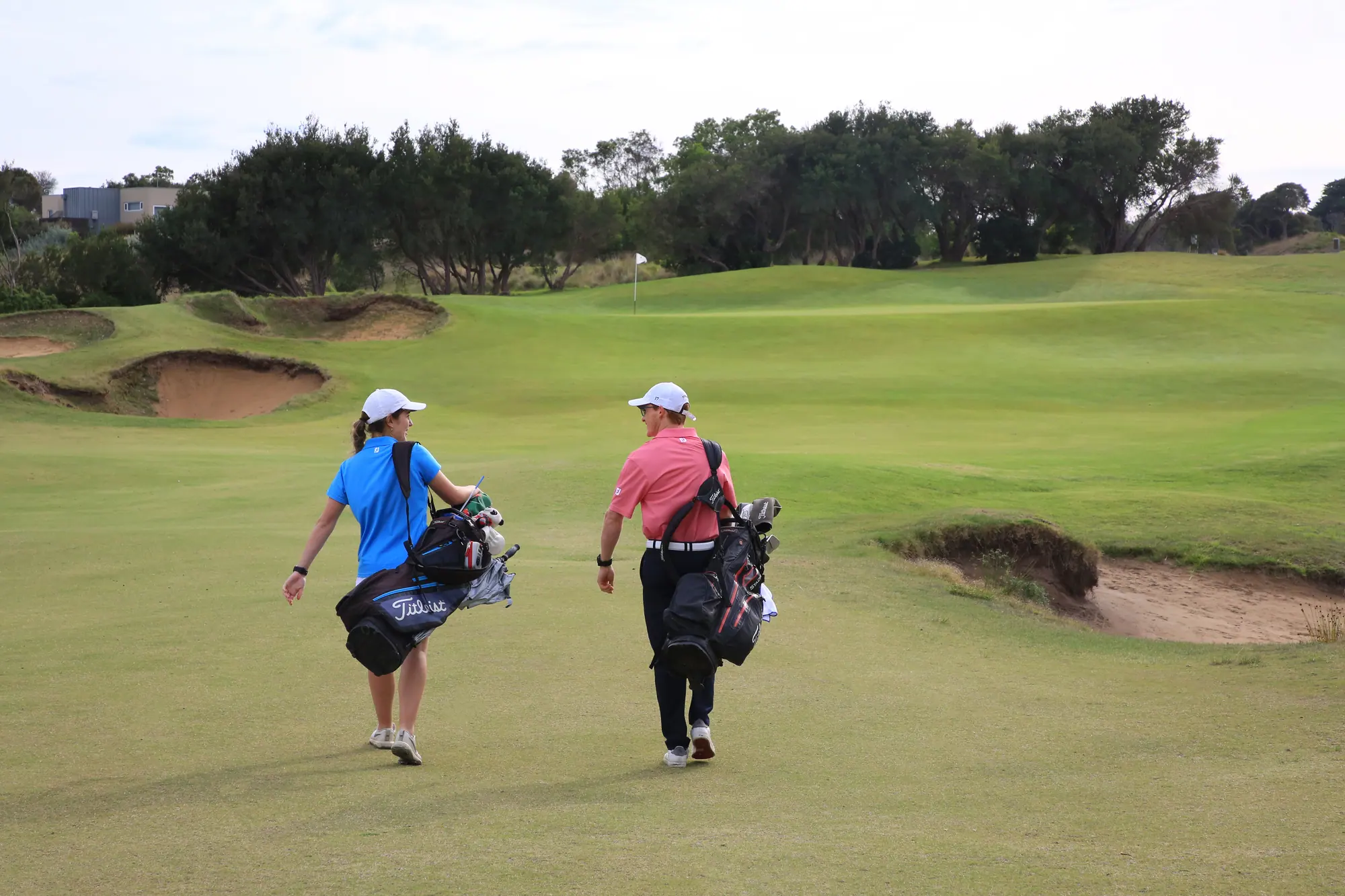 Two golfers carry their bags along a fairway at Moonah Links between deep sand bunkers towards a distant flag