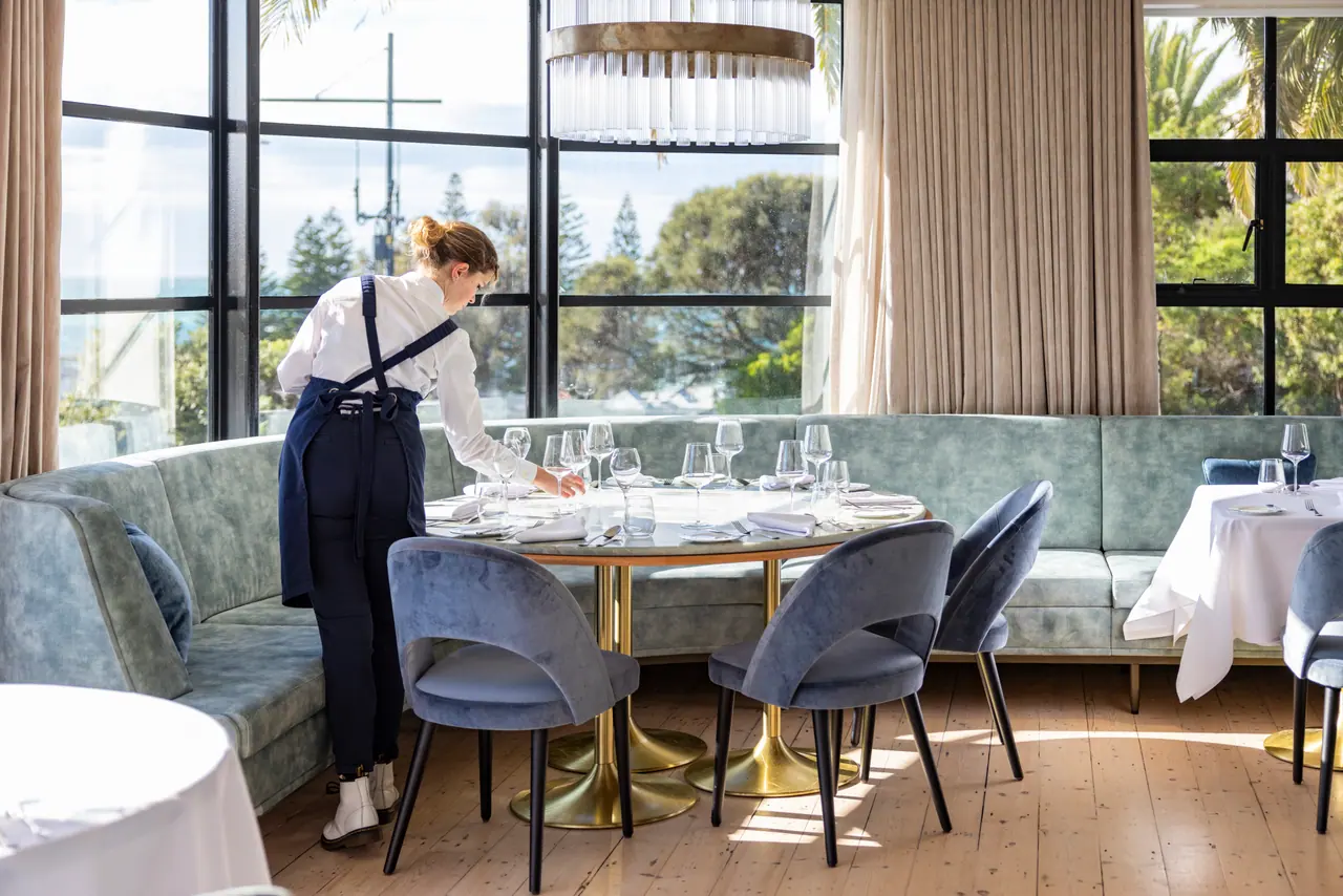 A staff member sets glasses on a round table at Audrey's, The Continental Sorrento, beside a velvet banquette and tall windows