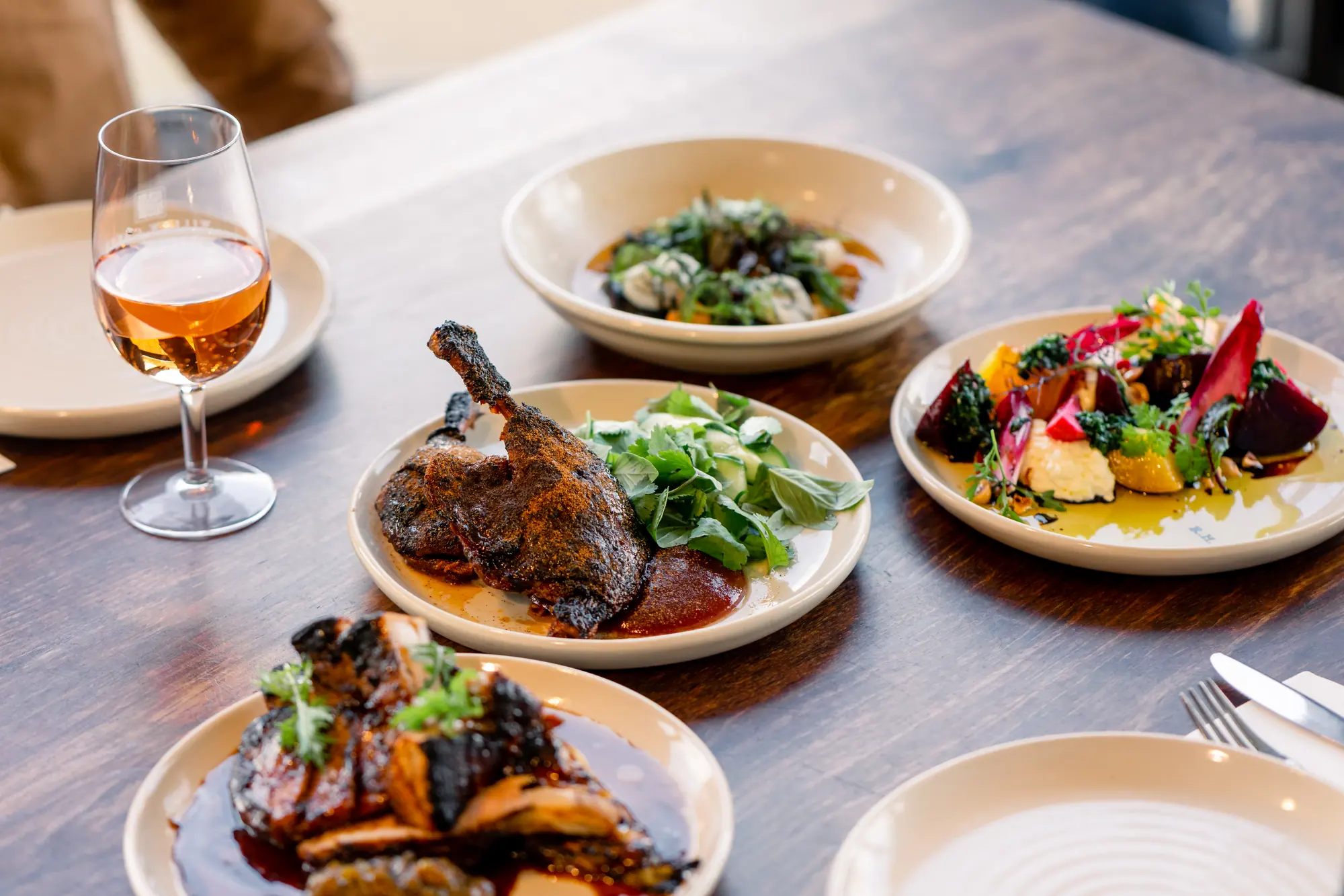 Plates of roast duck, charred meat, a beetroot salad and a leafy dish with a glass of rose on a timber table