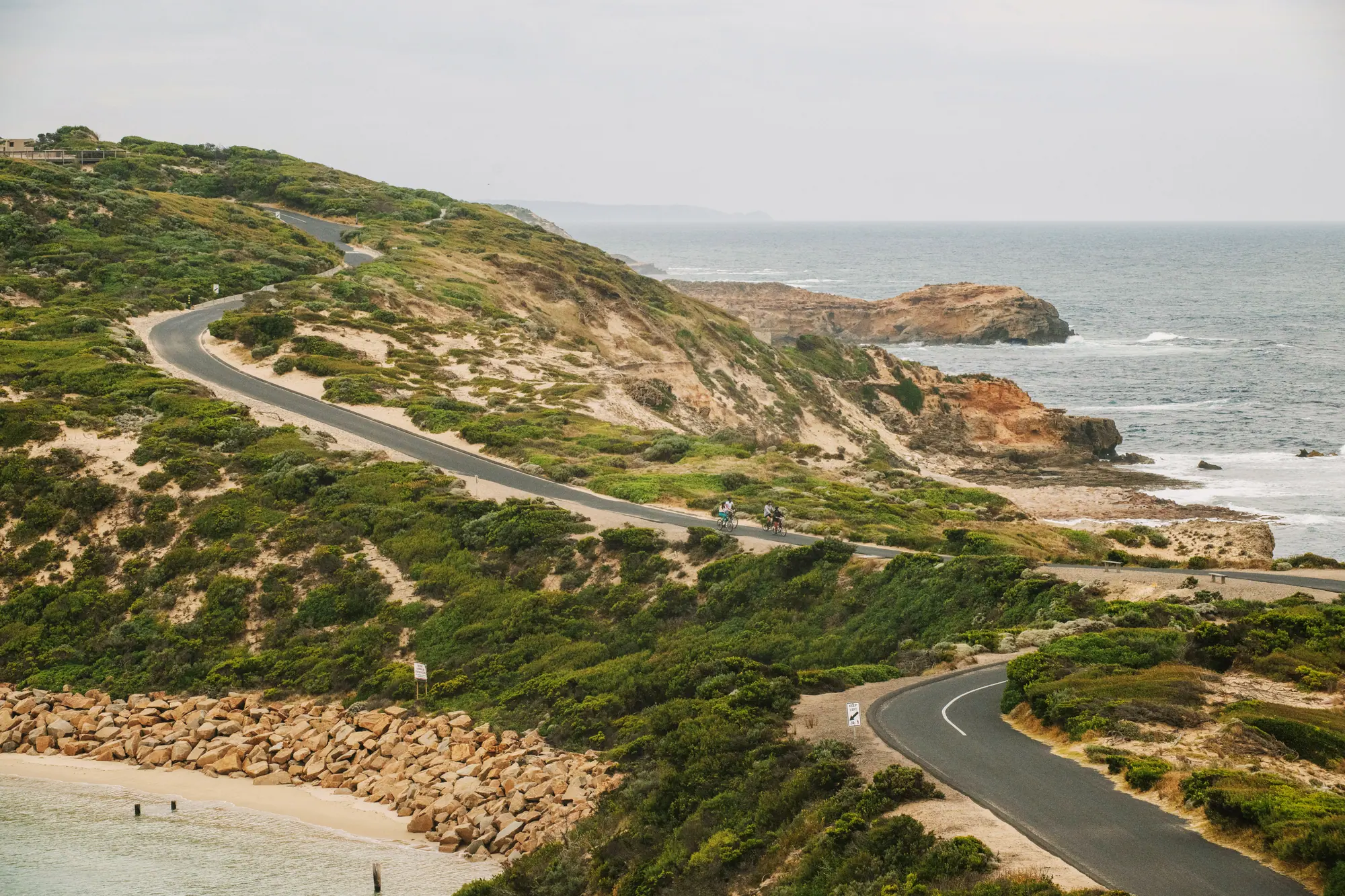 Two cyclists on a road winding through coastal scrub and sandy dunes at Point Nepean above rocky headlands and a grey sea