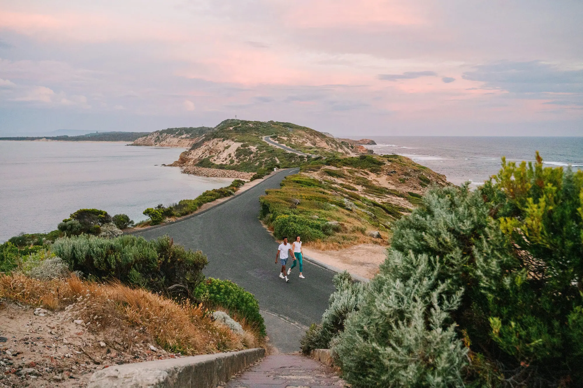 Two walkers on the sealed road to Fort Nepean as it winds along the narrow headland between bay and ocean under a pink sky