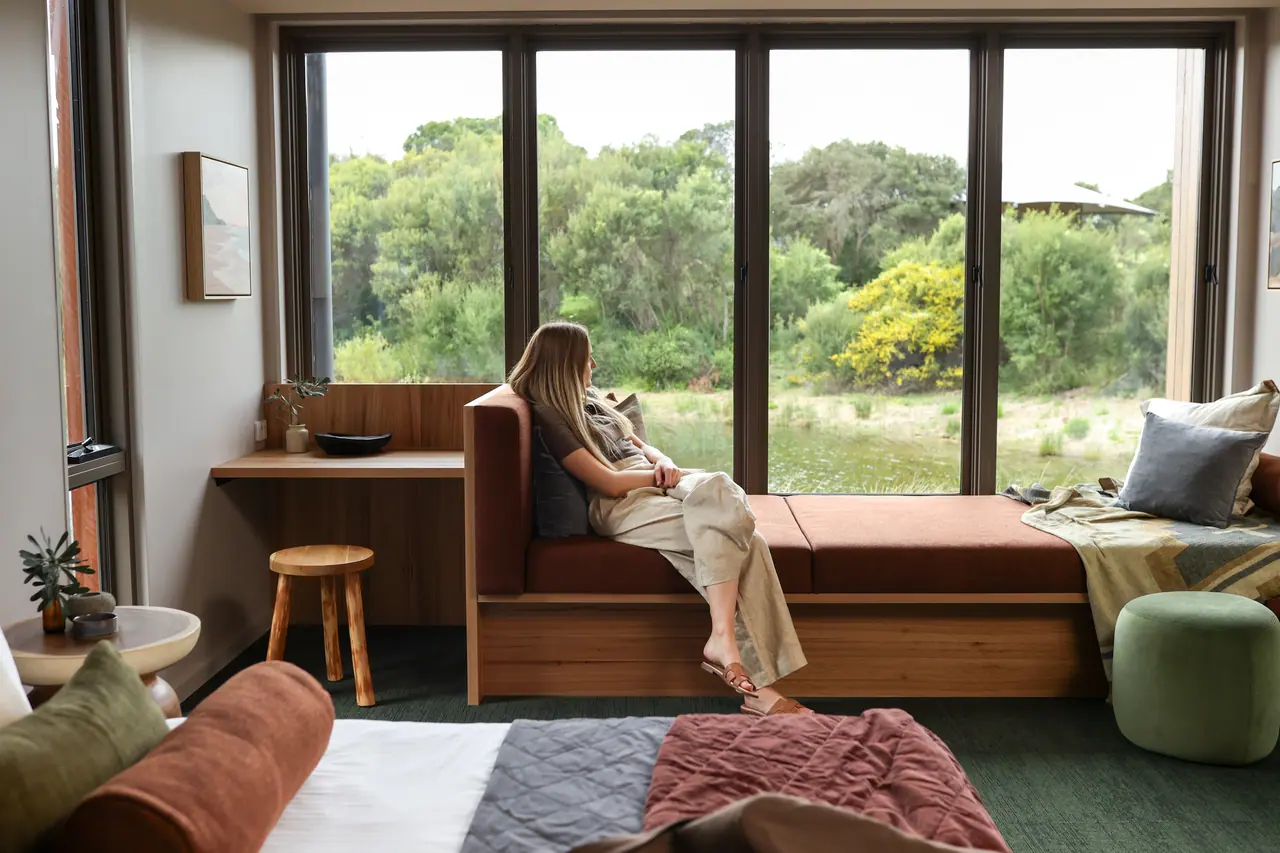 A guest sits beside a bushland-view window in a Peninsula Hot Springs Eco Lodge room.